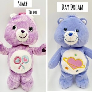 May include: Two plush bear toys. The left bear is purple tie-dye with a white belly patch featuring two crossed lollipops and the text SHARE TIE DYE. The right bear is solid periwinkle blue with a white belly patch showing a pink heart with a ring and stars, labeled DAY DREAM.