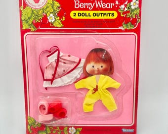 May include: Vintage Strawberry Shortcake Berry Wear doll outfit set. The packaging is red with the words "Strawberry Shortcake Berry Wear" in white and yellow. The set includes a doll, a pink dress, yellow jumpsuit, and red shoes.