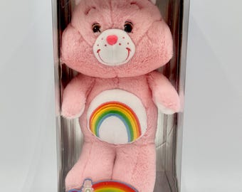 May include: A pink Cheer Bear plush toy in a collector's edition box. The bear has a rainbow on its belly and a smiling face with a red nose. The box has the Care Bears logo and the words "Cheer Bear".