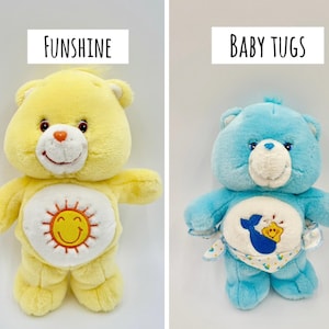 May include: Two plush bear toys. The left bear is yellow with a white belly patch featuring an orange sun with a smiling face and the text FUNSHINE above it. The right bear is light blue with a white belly patch showing a blue whale and a yellow star, wearing a patterned diaper with the text BABY TUGS above it.