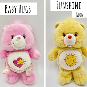 May include: Two plush bear toys. The left bear is pink with a white belly patch featuring a yellow star inside a pink heart and the text Baby Hugs. The right bear is yellow with a white belly patch featuring a yellow sun and the text Funshine Glow.