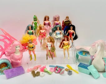 May include: A collection of vintage fashion dolls with various hairstyles and outfits, including superhero and fantasy themes. Accessories like brushes, shields, and wings are also present, along with a pink decorative display.