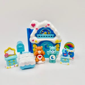 May include: A collection of Care Bears toys, including a house, two bears, a bed, a dresser, and a rainbow. The house is blue with a white cloud roof and the words "Welcome Bedtime Bear" on it. The bears are orange and blue.