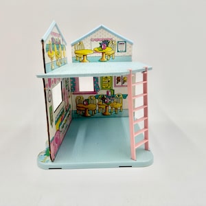 May include: A miniature, two-story dollhouse with a light blue exterior and a pink ladder. The interior features cartoon illustrations of tables, chairs, and food items. The roof is light blue with a white trim.