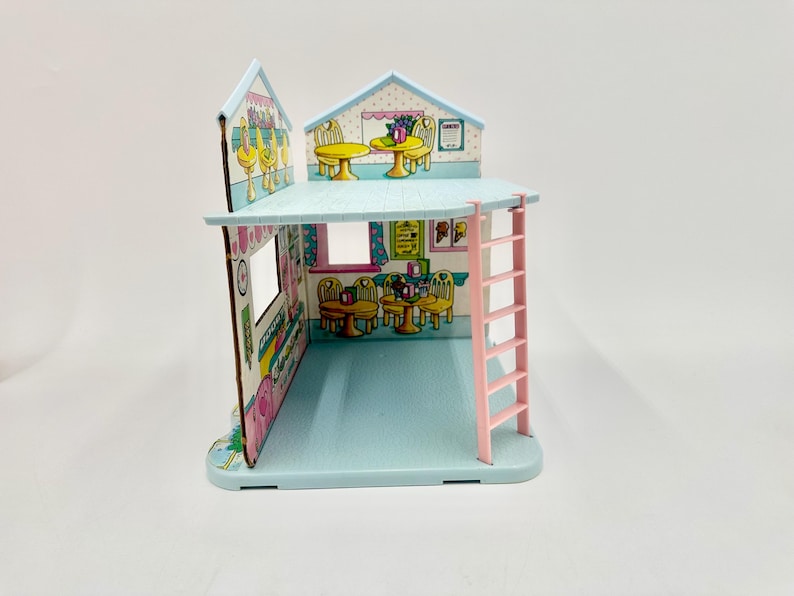 May include: A miniature, two-story dollhouse with a light blue exterior and a pink ladder. The interior features cartoon illustrations of tables, chairs, and food items. The roof is light blue with a white trim.
