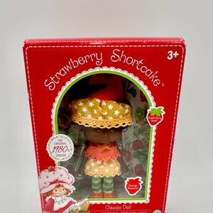 Pode incluir: Uma boneca clássica da Moranguinho numa caixa vermelha. A boneca tem um vestido amarelo e laranja, um chapéu a condizer e meias às riscas verdes e brancas. A caixa diz "Strawberry Shortcake" e "The Original 1980 Design!"