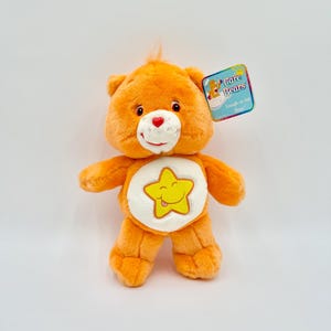 May include: An orange plush bear toy with a white circular patch on its chest featuring a yellow smiling star with its tongue sticking out. The bear has a red heart-shaped nose and a tag attached to its ear that reads Care Bears Laugh-a-lot Bear.