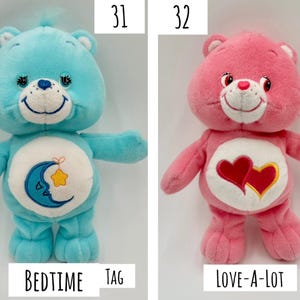 May include: Two plush Care Bears toys labeled 31 and 32. Number 31 is a light blue bear with a blue moon and yellow star emblem on its white belly and a Care Bears tag. Number 32 is a pink bear with two red hearts on its white belly labeled Love-A-Lot.
