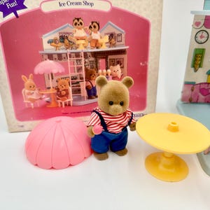 May include: A vintage Ice Cream Shop playset featuring a brown bear figure wearing a red and white striped shirt and blue overalls. The set includes a pink umbrella, yellow table, and a miniature ice cream shop with animal figurines.