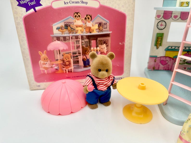 May include: A vintage Ice Cream Shop playset featuring a brown bear figure wearing a red and white striped shirt and blue overalls. The set includes a pink umbrella, yellow table, and a miniature ice cream shop with animal figurines.