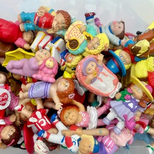 May include: A collection of vintage Cabbage Patch Kids figurines in various poses and outfits. The dolls are in a variety of colors, including pink, blue, yellow, and red. Some dolls are holding accessories like hearts and books.