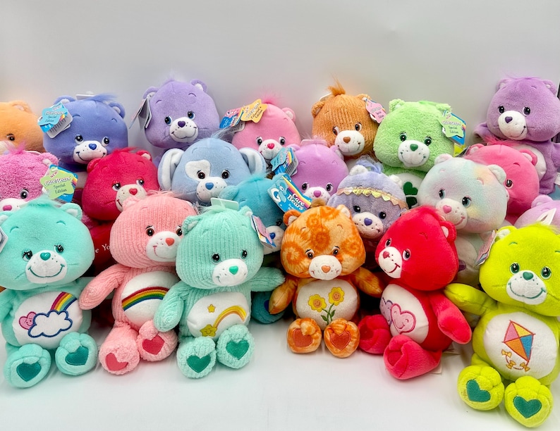 May include: A collection of various Care Bears plush toys in multiple colors including pink, blue, green, orange, purple, and red. Each stuffed animal features a unique embroidered belly badge, plastic nose, and attached product tags.