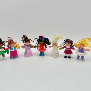 Op de afbeelding: Een verzameling van zeven kleine poppen met verschillende kapsels en outfits. De poppen hebben verschillende huidtinten en dragen jurken in kleuren als groen, roze, rood en wit. Sommige poppen houden kleine accessoires vast.