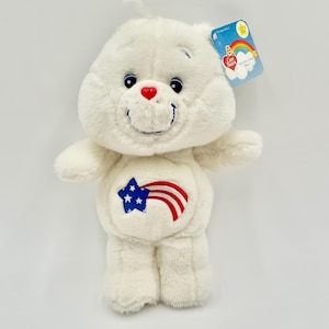 Könnte beinhalten: Ein weißes Care Bear Plüschtier mit roter Herz-Nase, blauen Augen und einem lächelnden Gesicht. Der Bär hat ein Stern- und Streifen-Design auf dem Bauch. Ein Anhänger mit einem Regenbogen und dem Care Bears-Logo ist angebracht.