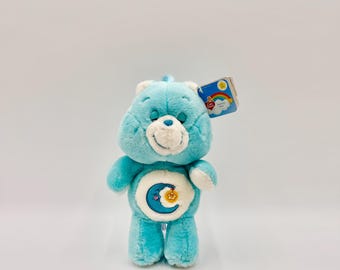 Könnte beinhalten: Ein hellblauer Plüschbär mit weißer Schnauze und einem runden Bauchaufnäher, der einen blauen Halbmond und einen gelben Stern zeigt. Der Bär hat geschlossene Augen und ein kleines Etikett am Ohr mit Regenbogenmotiv und dem Text Care Bears.