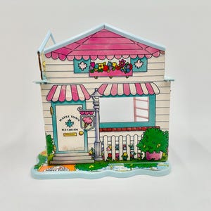 May include: A colorful Maple Town Ice Cream shop toy with a pink roof and awnings. The shop features a window, door, and a sign with an ice cream cone. The base is light blue with a decorative edge.