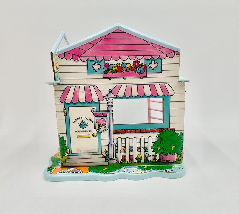 May include: A colorful Maple Town Ice Cream shop toy with a pink roof and awnings. The shop features a window, door, and a sign with an ice cream cone. The base is light blue with a decorative edge.