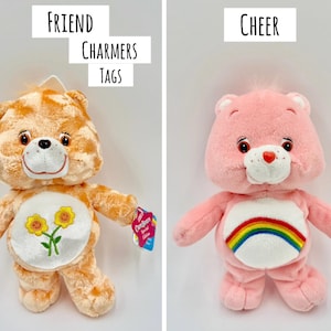 May include: Two plush bear toys. The left bear is orange with a white belly patch featuring two yellow flowers and the text "FRIEND CHARMERS TAGS" above it. The right bear is pink with a white belly patch featuring a rainbow and the text "CHEER" above it.