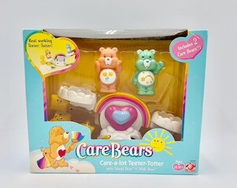 May include: A Care Bears Care-a-lot Teeter-Totter toy set in a box. The set includes two Care Bears, a rainbow, and other accessories. The box has the text "Real working Teeter-Totter!" and "Includes 2 Care Bears!"