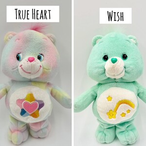 May include: Two plush bear toys. The left bear is multicolored with a pastel tie-dye pattern and a belly badge featuring a pink heart inside a star. The text above reads True Heart. The right bear is mint green with a shooting star belly badge. Text reads Wish.