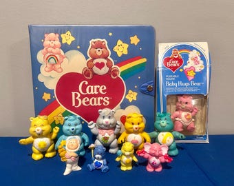 May include: A collection of Care Bears figurines, including a Baby Hugs Bear, a rainbow-colored book, and a pink and white box with the words "Care Bears" and "Baby Hugs Bear".