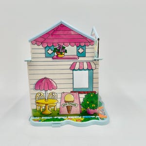 May include: A colorful, vintage ice cream shop playset. The playset features a pink roof, awning, and umbrella, with a sign advertising ice cream. The shop has a table and chairs, and a small garden with flowers.