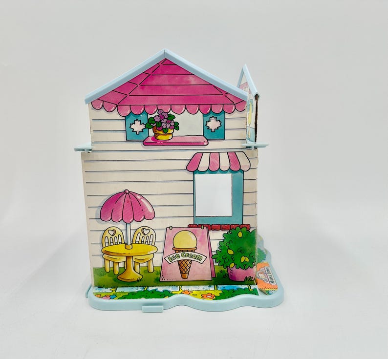 May include: A colorful, vintage ice cream shop playset. The playset features a pink roof, awning, and umbrella, with a sign advertising ice cream. The shop has a table and chairs, and a small garden with flowers.