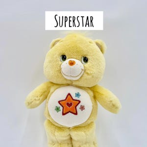 May include: A yellow plush bear with a white belly patch featuring an orange star with a red heart center, surrounded by smaller blue, green, and red stars. The text GLOW IN THE DARK and SUPERSTAR is printed above the bear on a white background.