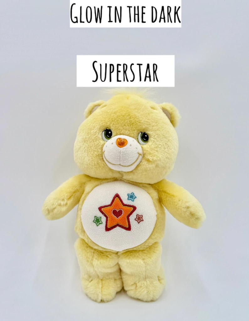 May include: A yellow plush bear with a white belly patch featuring an orange star with a red heart center, surrounded by smaller blue, green, and red stars. The text GLOW IN THE DARK and SUPERSTAR is printed above the bear on a white background.