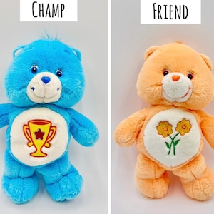May include: Two plush bears. The blue bear on the left is labeled CHAMP and features a yellow trophy emblem on its white belly patch. The orange bear on the right is labeled FRIEND and features two yellow flowers on its white belly patch.