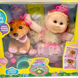 May include: Cabbage Patch Kids 35th Anniversary Deluxe Birthday Fashion set. Includes a doll with a pink dress and tiara, and a brown plush dog with a pink bow. The box has the text "It's Party Time!" and "We're Adoptable!"