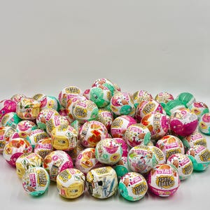 May include: A large collection of spherical Make It Mini Verse toy capsules featuring colorful graphic designs, various food illustrations, and branding text on their plastic surfaces, arranged in a dense pile against a plain background.