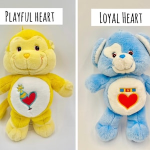 May include: Two plush toys: a yellow monkey labeled Playful Heart with a party hat and heart emblem on its belly, and a blue bear labeled Loyal Heart with a medal and heart emblem on its belly. Both have white belly patches and soft fur.