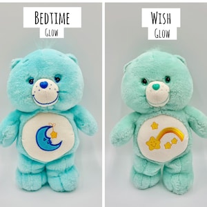 May include: Two plush bear toys with soft fur. The left bear is bright blue with a blue crescent moon and yellow star emblem, labeled BEDTIME GLOW. The right bear is mint green with a yellow rainbow and star emblem, labeled WISH GLOW.