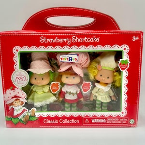 Puede incluir: Un juego de cajas rojas de muñecas Strawberry Shortcake de la década de 1980. La caja presenta un asa y una ventana transparente que muestra tres muñecas con diferentes colores de cabello y atuendos. El texto "Strawberry Shortcake" y "Classic Collection" son visibles.