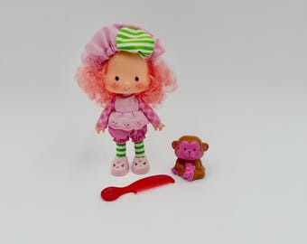 May include: A doll with pink curly hair wearing a pink bonnet with green and white stripes, a pink checkered top, and green and white striped leggings. Beside the doll is a small brown monkey figurine and a red plastic hair comb.