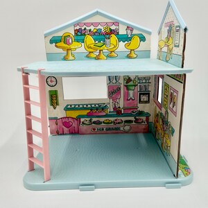 May include: A vintage, pastel-colored ice cream shop playset. The two-story structure features a pink ladder, a blue base, and printed interior details including tables, chairs, and an ice cream counter. The shop has a clock and the words "Ice Cream" on the counter.