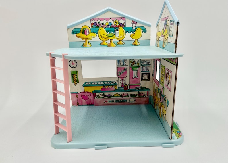 May include: A vintage, pastel-colored ice cream shop playset. The two-story structure features a pink ladder, a blue base, and printed interior details including tables, chairs, and an ice cream counter. The shop has a clock and the words "Ice Cream" on the counter.
