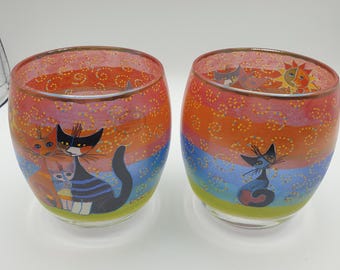 May include: Two rounded glass votive holders featuring hand-painted cat illustrations against a vibrant rainbow-striped background. The design includes swirling yellow patterns, a sun motif, and a metallic gold rim around the top edge of each glass.