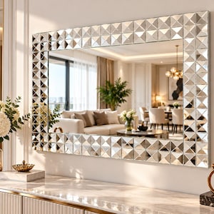 Könnte beinhalten: Ein rechteckiger Wandspiegel mit einem breiten Rahmen, der aus zahlreichen kleinen, reflektierenden, pyramidenförmigen Glasfliesen besteht. Die Spiegelfläche ist klar und glatt und spiegelt ein helles Wohnzimmer.