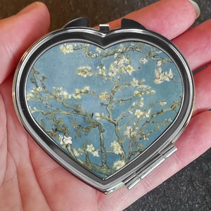 Könnte beinhalten: Ein silberner, herzförmiger Kompakt-Spiegel mit einem blauen und weißen Blumendesign. Das Design zeigt einen Baum mit weißen Blüten vor blauem Hintergrund.