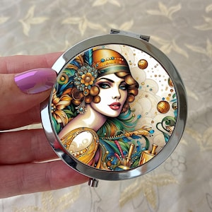 Peut inclure: Un miroir de poche rond et argenté avec une illustration art déco d'une femme aux yeux bleus, portant un chapeau et des bijoux. L'œuvre d'art comprend des nuances d'or, de sarcelle et d'orange. Le miroir est tenu dans une main avec du vernis à ongles rose.