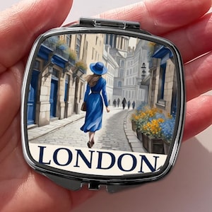 Op de afbeelding: Een zilveren compact spiegel met een kleurrijk beeld van een vrouw die door een straat in Londen loopt. Het beeld heeft de tekst "LONDON" onderaan.