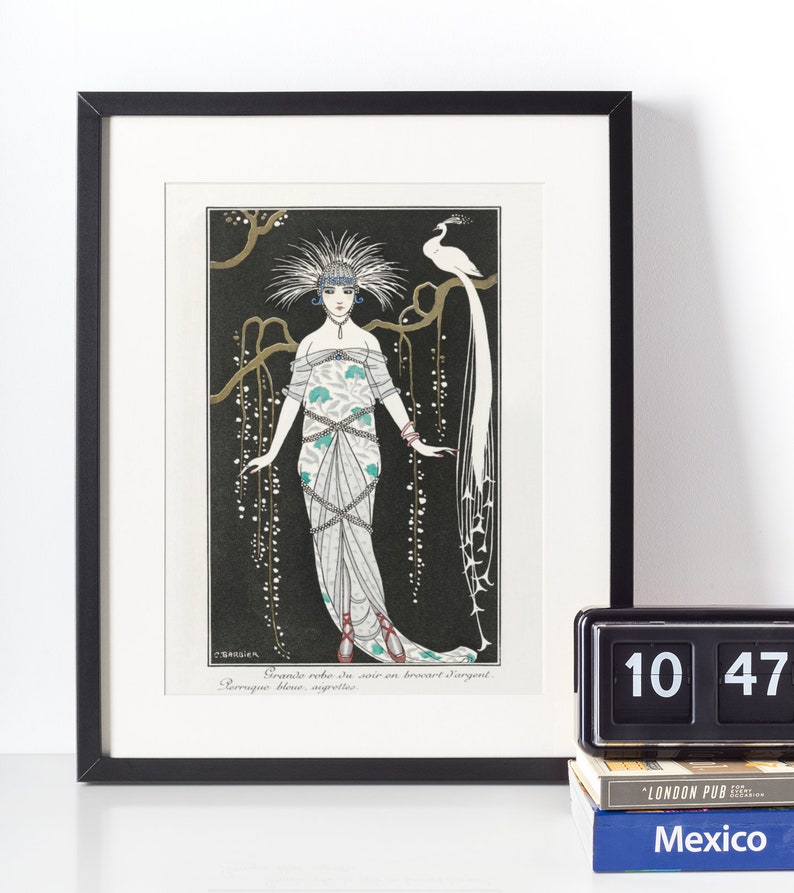 May include: Framed art print featuring an Art Deco illustration of a woman in an elaborate gown with a peacock. The artwork is in black, white, and teal, with the text "Grande robe du soir en brocart d'argent" and "Perruque bleue, angrelles".