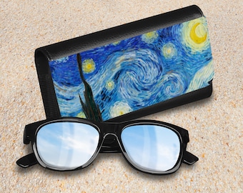 May include: A black sunglasses case with a print of Vincent van Gogh's "Starry Night" painting. The case is lying on a sandy surface next to a pair of black sunglasses.
