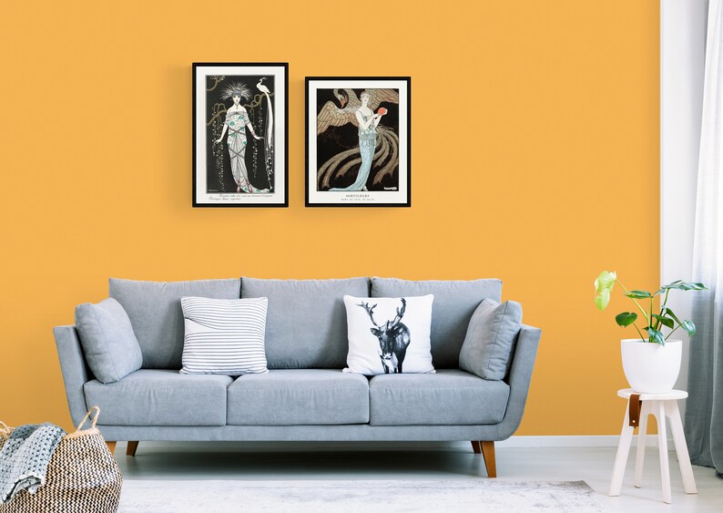 May include: Two framed art prints hang above a gray sofa against a yellow wall. The prints feature Art Nouveau illustrations of women with ornate details and dark backgrounds. A striped pillow and a deer pillow decorate the sofa.
