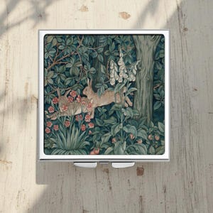 Op de afbeelding: Een vierkante, zilverkleurige compacte spiegel met een bosscène met twee konijnen. Het kunstwerk toont een weelderig bos met groene bladeren, rode bloemen en witte vingerhoeden. De compact heeft een scharnierende opening en een reflecterend oppervlak.