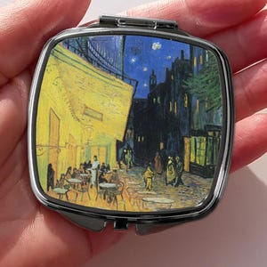 以下が含まれることがあります： ゴッホの「夜のカフェテラス」のプリントが施された、正方形のシルバーフレームのコンパクトミラー。このアートワークは、客がいる黄色いカフェ、暗い通り、星空を描いています。鏡は手で持たれています。