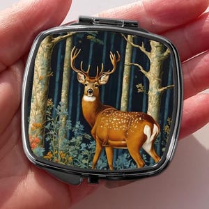 May include: A compact mirror with a silver metal frame and a colorful image of a deer in a forest setting.