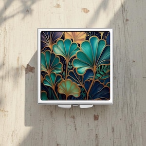 Könnte beinhalten: Ein quadratischer, silberner Kompaktspiegel mit einem teal- und goldfarbenen Blumendesign. Das vom Art-déco inspirierte Muster umfasst stilisierte Blätter und Blüten in Teal, Gold und Marineblau vor einem dunklen Hintergrund. Der Kompaktspiegel liegt auf einer hellen Holzoberfläche.